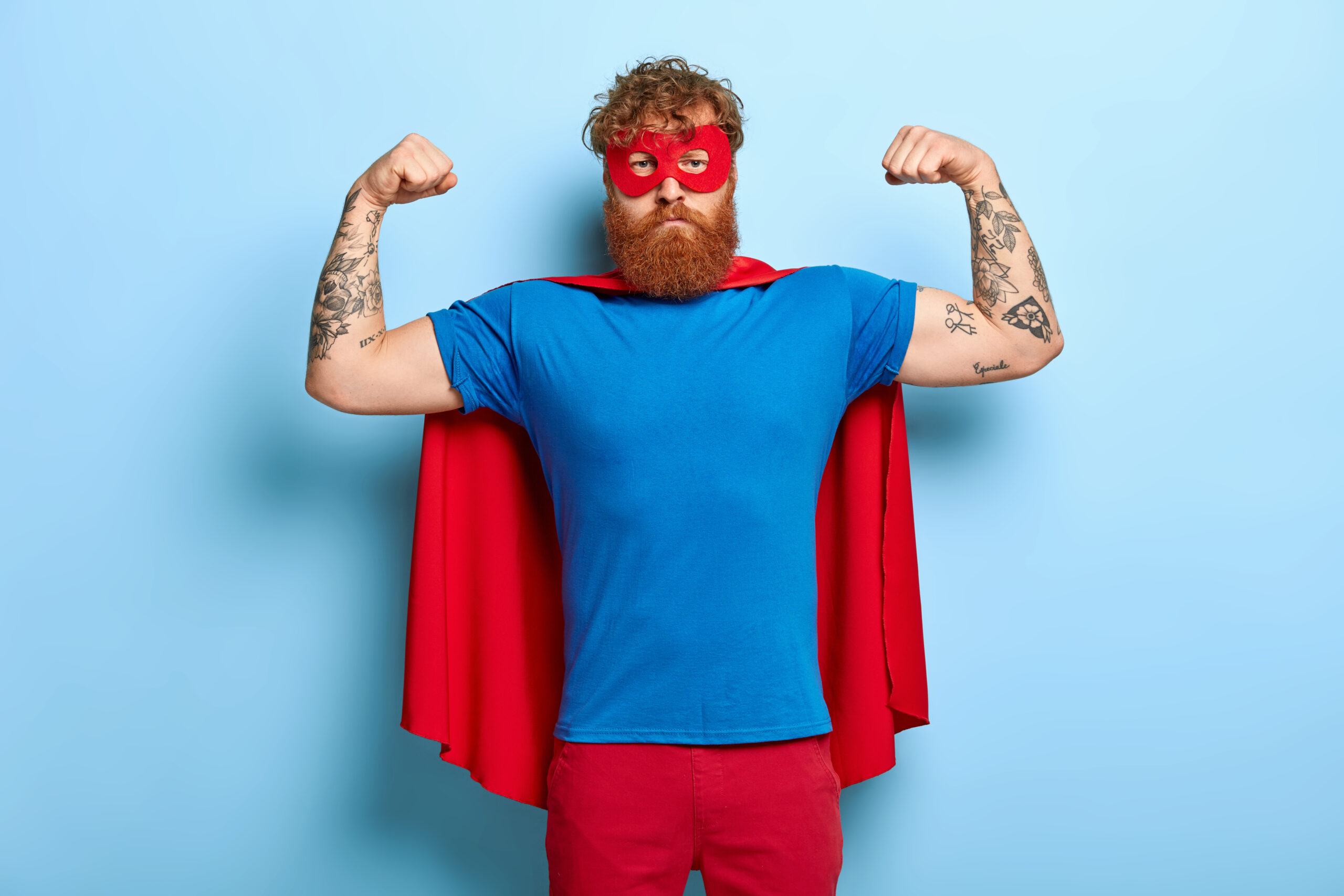 Successful hero wears red mask and cape, raises arms, shows biceps, demonstrates courage and strength, looks serious and confident, poses against blue wall. Real superhero ready to help you.